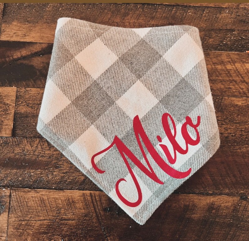 May include: A gray and white plaid dog bandana with the name "Milo" in red script. The bandana is a triangle shape and is made of a soft fabric. The background is a wooden surface.