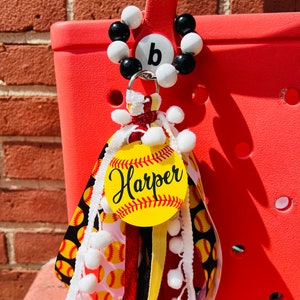 K&ouml;nnte beinhalten: Ein gelber und schwarzer Softball-Anh&auml;nger mit dem Namen "Harper" darauf. Der Anh&auml;nger ist an einem roten, schwarzen und gelben Band mit wei&szlig;en Pompons befestigt. Der Anh&auml;nger ist an einem schwarzen und wei&szlig;en Perlenanh&auml;nger mit einem Buchstaben "b" befestigt.