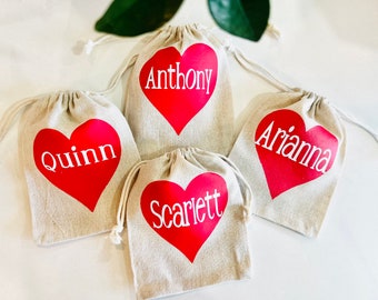 May include: Four beige drawstring bags with red heart designs. Each heart has a name printed in white: Quinn, Anthony, Arianna, and Scarlett. The bags are made of a natural-colored fabric.