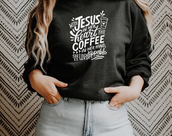 May include: Black sweatshirt with white text: "With Jesus in her heart and coffee in her hand, she is unstoppable." The design includes a coffee cup graphic. The person is wearing light blue jeans.
