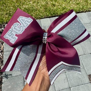 May include: A large maroon cheerleading bow with silver glitter accents and the letters "FCA" in silver glitter. The bow has a rhinestone center and white stripes. The bow is made of ribbon.
