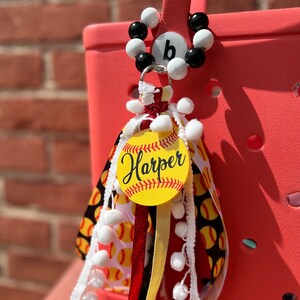 K&ouml;nnte beinhalten: Ein roter und wei&szlig;er Softball-Taschenanh&auml;nger mit einem wei&szlig;en und schwarzen Perlenanh&auml;nger mit dem Buchstaben "b" darauf. Der Anh&auml;nger zeigt einen gelben Softball mit dem Namen "Harper" darauf. Der Anh&auml;nger ist mit wei&szlig;en Pompons und roten, wei&szlig;en und schwarzen B&auml;ndern verziert.