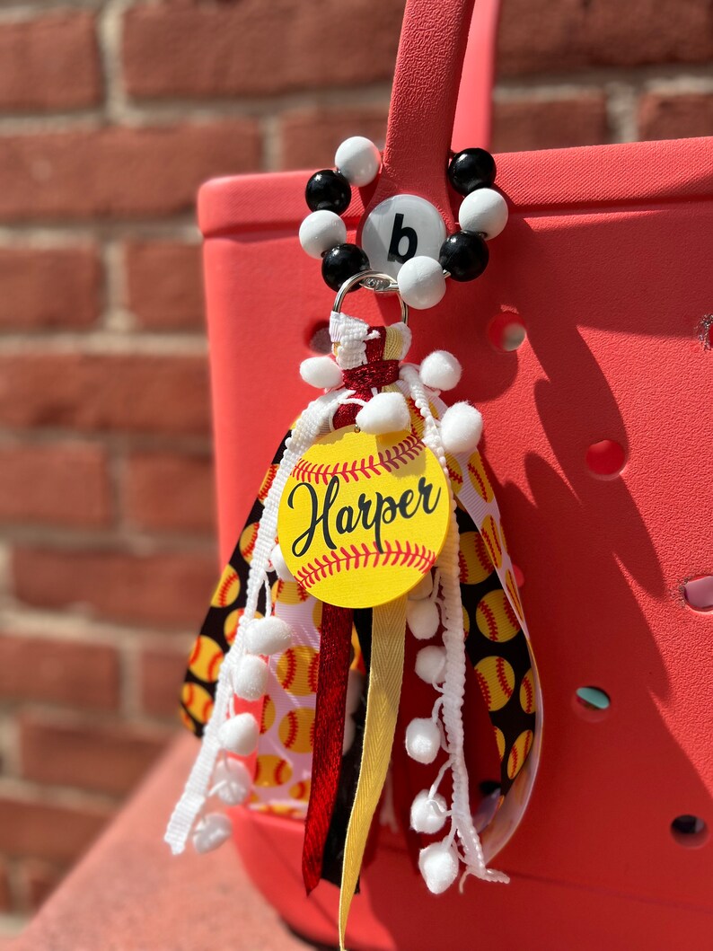K&ouml;nnte beinhalten: Ein roter und wei&szlig;er Softball-Taschenanh&auml;nger mit einem wei&szlig;en und schwarzen Perlenanh&auml;nger mit dem Buchstaben "b" darauf. Der Anh&auml;nger zeigt einen gelben Softball mit dem Namen "Harper" darauf. Der Anh&auml;nger ist mit wei&szlig;en Pompons und roten, wei&szlig;en und schwarzen B&auml;ndern verziert.