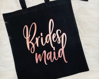 May include: Black tote bag with rose gold lettering that reads "Bridesmaid". The bag has two handles and is made of a sturdy fabric. The bag is sitting on a white, fluffy surface.