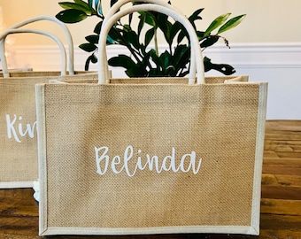 Può includere: Borsa tote in iuta beige con finiture e manici bianchi. Il nome "Belinda" è stampato in bianco sul davanti. Una seconda borsa con il nome "Kim" è parzialmente visibile. Fogliame verde sullo sfondo.