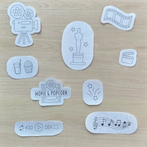 May include: A set of nine black and white movie themed iron-on patches. The patches include a film reel, a movie camera, a popcorn bucket, a glass of water, a clapperboard, a film strip, a music note, a star, and a play button.