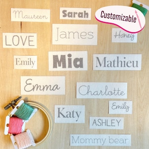 Puede incluir: Una colección de etiquetas de tela rectangulares con varios nombres y palabras como Maureen, Sarah, James, Honey, LOVE, Emily, Mia, Mathieu, Emma, Charlotte, Katy, ASHLEY y Mommy bear, junto a un bastidor de bordado y tres carretes de hilo.