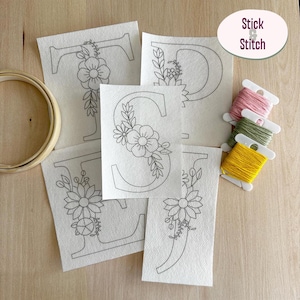 Op de afbeelding: Vijf witte stoffen vierkanten met zwarte lijntekeningen van de letters T, P, S, I en J, versierd met bloemmotieven. Een houten borduurring en drie kleine plastic klosjes met roze, groen en geel borduurgaren zijn inbegrepen. De tekst Stick & Stitch is zichtbaar.
