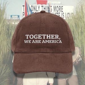 May include: A brown corduroy baseball cap with white embroidered text reading "TOGETHER, WE ARE AMERICA". The cap features a curved peak and a rounded crown. The background includes a sign that says "ONLY THING MORE HATE IS LOVE".