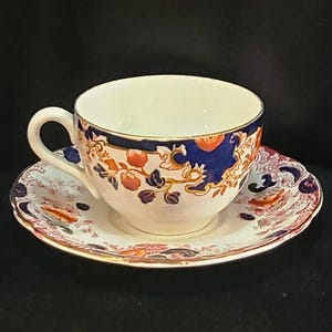 May include: A white teacup and saucer set with a detailed floral design in shades of blue, orange, and gold. The cup has a curved handle and a blue rim. The saucer has a scalloped edge. The set is likely vintage or antique.