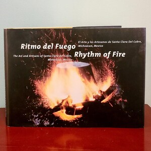 May include: A book titled "Ritmo del Fuego" with the English translation "Rhythm of Fire." The cover features a fiery image of a burning object. Text on the cover indicates the book is about the art and artisans of Santa Clara del Cobre, Michoacán, Mexico.