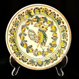 May include: A hand-painted ceramic plate featuring a rooster design in the center, surrounded by a wreath of yellow flowers and green leaves. The plate has a cream-colored background with a decorative border of yellow and green patterns. The plate is displayed on a clear stand.