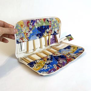 Peut inclure: Étui à aiguilles à tricoter crème ouvert, avec un motif patchwork coloré. L'étui est rempli d'aiguilles à tricoter en bois et d'une jauge violette. Le patchwork présente des motifs floraux bleus, jaunes et blancs.