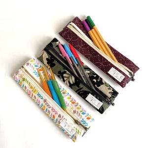 Op de afbeelding: Drie stoffen etuis met ritssluiting, elk gevuld met pennen of haaknaalden. Eén etui heeft een bloemenpatroon, één camouflage en de derde is bordeauxrood met witte stiksels. De etuis zijn open en tonen hun inhoud.