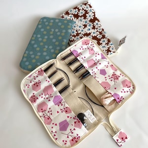 May include: A white canvas roll-up case with a zipper closure and a pink and white geometric pattern. The case is open and contains a set of knitting needles and a tape measure. The case is lined with a brown fabric with a white floral pattern.