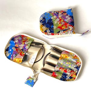 Puede incluir: Un estuche de agujas de tejer abierto y colorido y una bolsa con cremallera a juego. El estuche es blanco con un diseño de patchwork de patrones florales y geométricos en rojo, azul, verde y amarillo. Dentro del estuche hay agujas de tejer y accesorios.