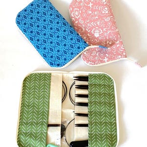 May include: A green and white chevron patterned knitting needle case, open to reveal needles and accessories. Two closed cases are visible: a blue patterned case and a pink floral case. The cases are for knitting.