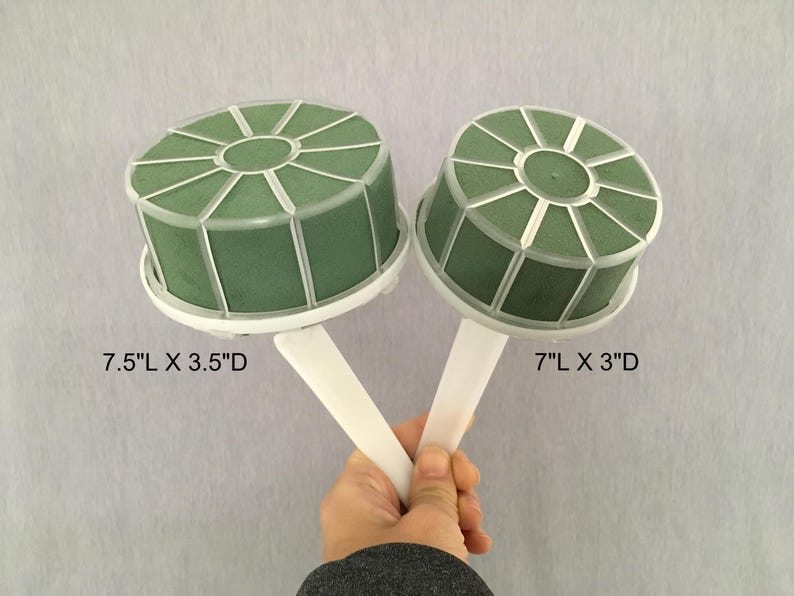 May include: Two round green floral foam cages with white plastic frames and handles. The left cage is labeled 7.5"L x 3.5"D and the right cage is labeled 7"L x 3"D. These floral foam holders are designed for arranging flowers and greenery.