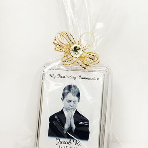 May include: A clear plastic bag with a gold ribbon and a clear plastic frame with a black and white photo of a boy in a suit praying. The frame has the text "My First Holy Communion" and "Jacob R. 3-27-2014".