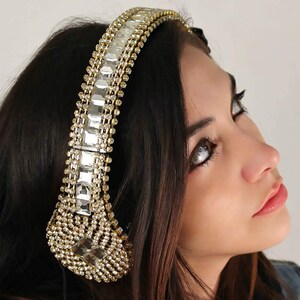 Puede incluir: Auriculares de diadema con una estructura en tono dorado incrustada con pedrer&iacute;a transparente rectangular y redonda. La diadema y las copas est&aacute;n completamente cubiertas de cristales brillantes, creando una superficie met&aacute;lica reflectante.