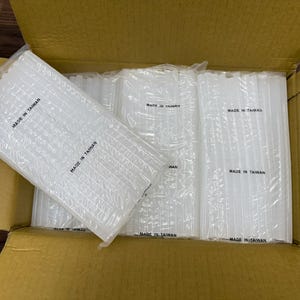 May include: Multiple bundles of clear, cylindrical hot glue sticks are arranged inside a cardboard box. Each individual plastic-wrapped package features the text MADE IN TAIWAN printed in black ink on the surface of the clear plastic packaging.