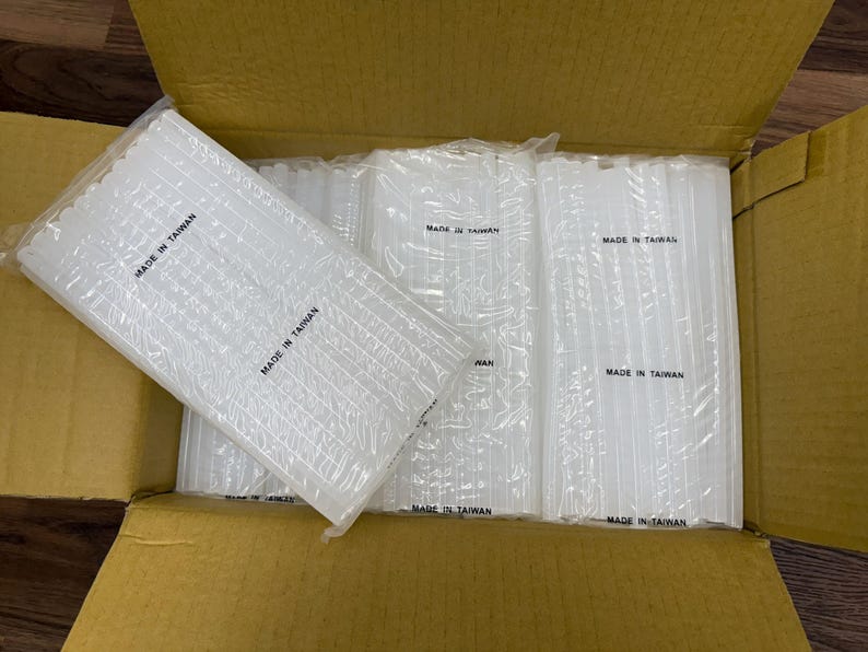 May include: Multiple bundles of clear, cylindrical hot glue sticks are arranged inside a cardboard box. Each individual plastic-wrapped package features the text MADE IN TAIWAN printed in black ink on the surface of the clear plastic packaging.