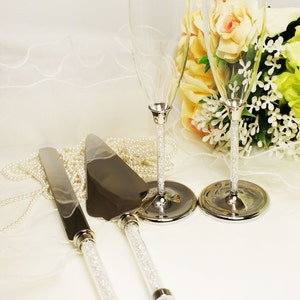 May include: A wedding cake cutting set with two champagne flutes and a knife and server. The flutes and handles of the knife and server are decorated with rhinestones.