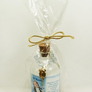 May include: A small glass bottle with a cork stopper, filled with a clear liquid and decorated with a blue rosary and a gold charm. The bottle has a label with a photo of a young boy, the text "Mi Bautiz Lou 2-15-2015", and a Spanish prayer.