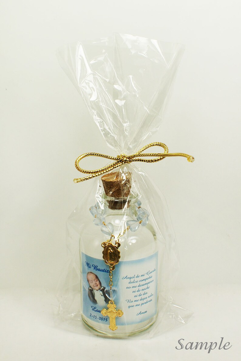 May include: A small glass bottle with a cork stopper, filled with a clear liquid and decorated with a blue rosary and a gold charm. The bottle has a label with a photo of a young boy, the text "Mi Bautiz Lou 2-15-2015", and a Spanish prayer.