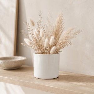 May include: A white cylindrical ceramic vase filled with a dried floral arrangement consisting of beige pampas grass, fluffy bunny tail grass, and delicate sprigs of small, light-colored dried foliage, creating a neutral and textured display.