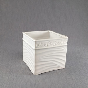 May include: A square, white ceramic planter with a decorative, raised border. The planter's sides feature a textured, wave-like pattern. The top edge has a detailed, embossed design. The planter is empty and sits on a grey surface.