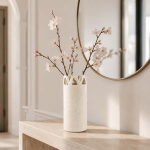 May include: A cylindrical off-white vase with a textured surface and a scalloped, open-work rim. The vase holds several slender branches featuring small, delicate pale pink cherry blossom flowers with dark brown stems and unopened buds.