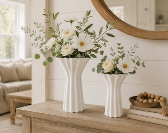 May include: Two white ceramic vases with a fluted, vertical ribbed design. The taller vase on the left holds a large arrangement of white cosmos flowers and green eucalyptus foliage. The shorter vase on the right holds a smaller floral arrangement.