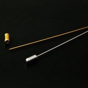 Könnte beinhalten: Zwei Nähnadeln, eine goldene und eine silberne, auf schwarzem Hintergrund. Die goldene Nadel hat einen runden Kopf und die silberne Nadel hat einen spitzen Kopf.