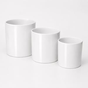 May include: Three cylindrical white ceramic containers of varying heights arranged in a descending row. Each vessel features a smooth, glossy finish, a flat base, and an open top with a simple, clean rim, presenting a uniform and minimalist design.