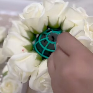May include: A circular arrangement of white artificial roses surrounding a central green plastic floral foam cage. The roses feature layered petals with green sepals at the base, creating a dense, rounded floral structure for decorative use.