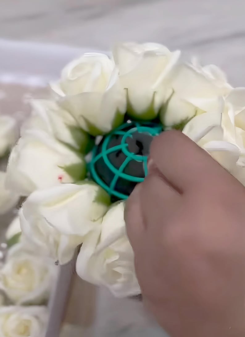 May include: A circular arrangement of white artificial roses surrounding a central green plastic floral foam cage. The roses feature layered petals with green sepals at the base, creating a dense, rounded floral structure for decorative use.