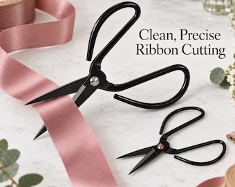 May include: Two pairs of black metal scissors with pointed blades and large looped handles, positioned next to a spool of dusty pink satin ribbon. The text Clean, Precise Ribbon Cutting is printed in black on the white surface near the scissors.