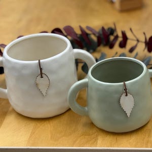 May include: Two ceramic mugs, one white and one sage green, with large handles. Each mug has a small, leaf-shaped wooden tag attached. The mugs are on a light-colored wooden surface, with dark red and green foliage in the background.
