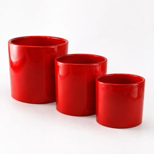 May include: A set of three cylindrical containers with a glossy, vibrant red finish. The containers are arranged in a descending order of size from left to right, each featuring a smooth, solid-colored exterior and a simple, open-top design.