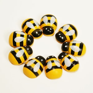 May include: A circle of nine wooden bee figurines, painted yellow with black stripes and white wings. The bees are arranged in a circle on a white background.