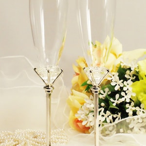 Könnte beinhalten: Zwei silberne Champagnerflöten mit kristallartigen Verzierungen an der Basis. Die Flöten stehen auf einer weißen Oberfläche mit einer Perlenkette und einem Strauß gelber und weißer Blumen.