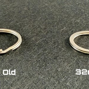 May include: Two silver-colored key rings, one labeled "32mm Old" and the other "32mm New", are displayed on a dark gray surface. The key rings are circular with a split for attaching keys. The text is white.