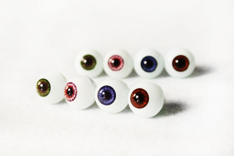 Puede incluir: Una fila de siete ojos de mu&ntilde;eca blancos con iris de diferentes colores. Los iris son verdes, rojos, rosas, azules, marrones, morados y rojos.