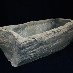 Puede incluir: Una jardinera rectangular gris, similar a la piedra, con una superficie rugosa y texturizada. La jardinera tiene un interior hueco y está diseñada para contener plantas. El fondo oscuro resalta la forma y la textura de la jardinera.