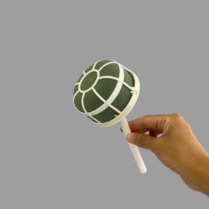 Puede incluir: Una bola de espuma floral blanca con un centro verde y un mango de plástico blanco. La bola está diseñada para sujetar flores y otros arreglos florales.