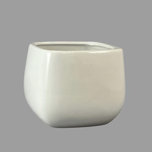 Puede incluir: Una maceta de cerámica blanca de forma cuadrada con esquinas redondeadas. Tiene un acabado liso y brillante y una amplia abertura. El diseño es minimalista y moderno, adecuado para varias plantas y estilos de decoración.
