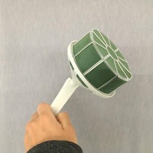 May include: A floral foam bouquet holder featuring a round, dark green foam block encased in a white plastic cage with a white handle. The structure is designed to support floral arrangements, providing a stable base for stems to be inserted.