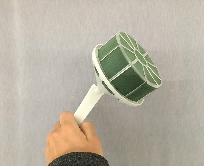 May include: A floral foam bouquet holder featuring a round, dark green foam block encased in a white plastic cage with a white handle. The structure is designed to support floral arrangements, providing a stable base for stems to be inserted.