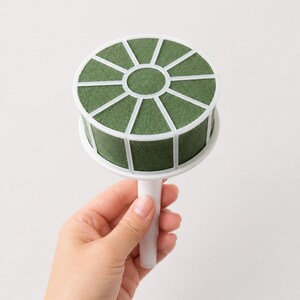 May include: A round floral foam cage with a white plastic frame and handle. The dark green foam is divided into segments by the white plastic grid, designed to hold flower stems securely in place for arrangements and floral displays.